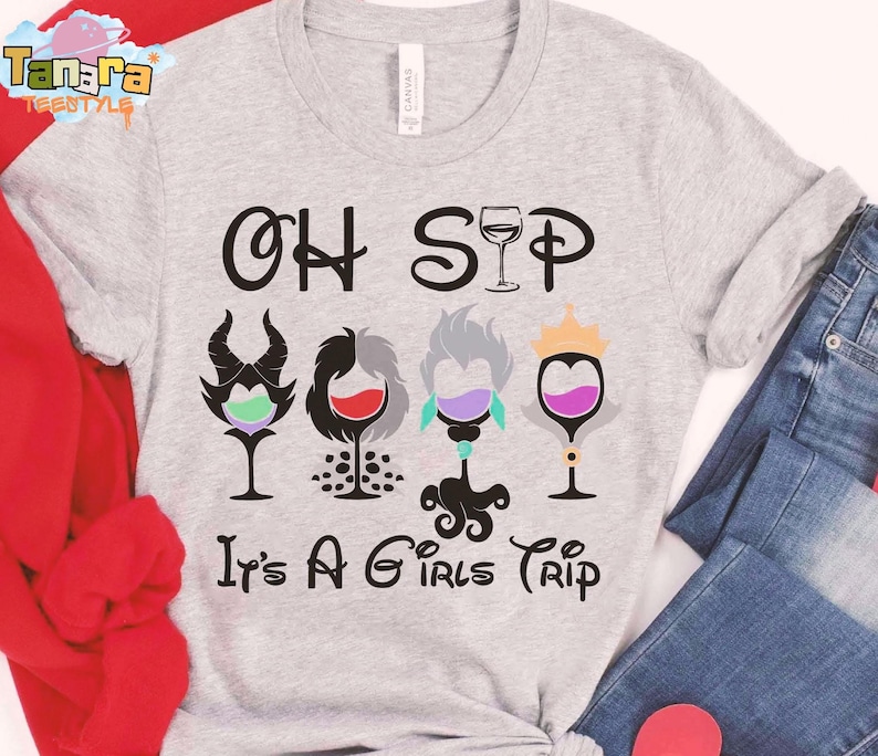 May include: Pale grey t-shirt with the words "OH SIP" and "It's A Girls Trip". The design shows four wine glasses with cartoon character-inspired designs, each filled with a different coloured liquid. The shirt is made of a soft material.