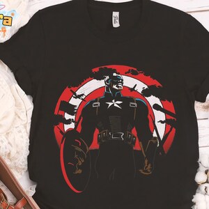 K&ouml;nnte beinhalten: Schwarzes T-Shirt mit Captain America-Grafik. Das Design zeigt einen Schild, einen Stern und den Text "Tanara Teenstyle". Die Grafik ist in Rot, Wei&szlig; und Blau gehalten.