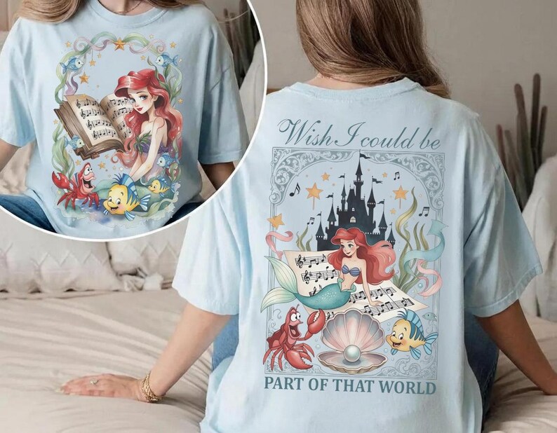 May include: Light blue t-shirt featuring a colorful illustration of a mermaid with red hair, a crab, and a fish. The front of the shirt shows the mermaid reading a book. The back of the shirt has the text "Wish I could be PART OF THAT WORLD".