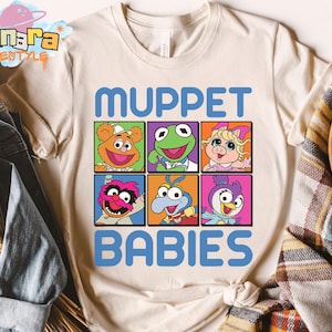 May include: Cream-colored t-shirt featuring the text "MUPPET BABIES" in blue, with a grid of six Muppet Babies character portraits. The portraits are in colorful squares, showcasing characters like Kermit and Miss Piggy.