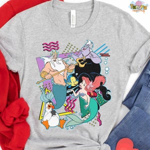 May include: A light grey t-shirt with a vibrant graphic of characters from The Little Mermaid. The design includes King Triton, Ariel, Flounder, Sebastian, Ursula, and Scuttle, set against a retro 80s style background.