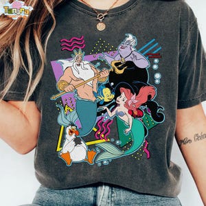 May include: Dark grey t-shirt with a vibrant graphic of characters from The Little Mermaid. The design includes King Triton, Ariel, Ursula, Flounder, and Scuttle, set against a retro 80s style background. The shirt has the Tanara logo in the top left corner.