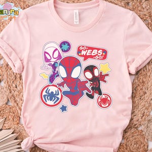 May include: A light pink t-shirt featuring a colorful graphic of Spider-Man characters. The design includes Spider-Gwen, Spider-Man, and Miles Morales, with the text "GO-WEBS-GO!" in a speech bubble. The shirt has rolled-up sleeves.