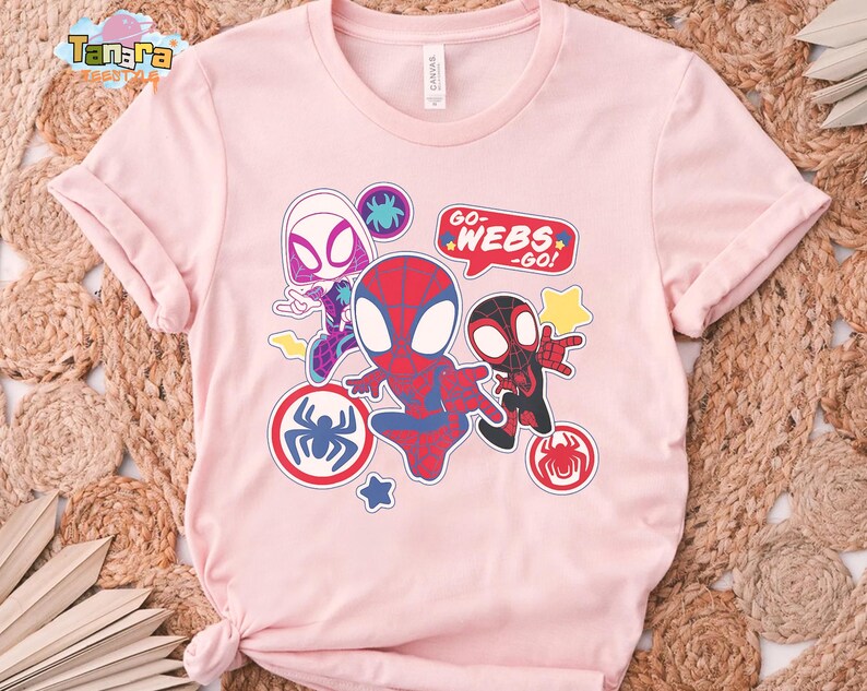 May include: A light pink t-shirt featuring a colorful graphic of Spider-Man characters. The design includes Spider-Gwen, Spider-Man, and Miles Morales, with the text "GO-WEBS-GO!" in a speech bubble. The shirt has rolled-up sleeves.