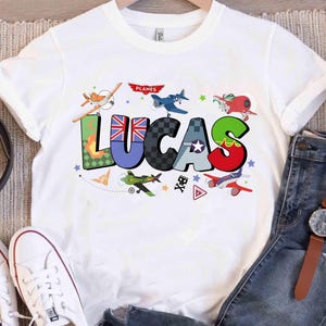 Op de afbeelding: Wit T-shirt met de naam "LUCAS" in kleurrijke, patroonletters en vliegtuigillustraties. De letter "U" heeft een Union Jack-ontwerp. Het woord "PLANES" staat boven de naam. Het shirt wordt gecombineerd met een jeans en witte sneakers.