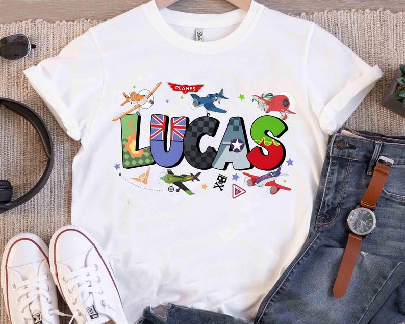 Op de afbeelding: Wit T-shirt met de naam "LUCAS" in kleurrijke, patroonletters en vliegtuigillustraties. De letter "U" heeft een Union Jack-ontwerp. Het woord "PLANES" staat boven de naam. Het shirt wordt gecombineerd met een jeans en witte sneakers.