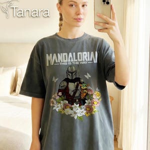 Puede incluir: Camiseta gris con el texto "MANDALORIAN - THIS IS THE WAY" y una ilustraci&oacute;n del Mandaloriano, Grogu y flores. La camiseta tiene mangas cortas y un corte holgado. La marca "Tanara" est&aacute; en la esquina superior izquierda.