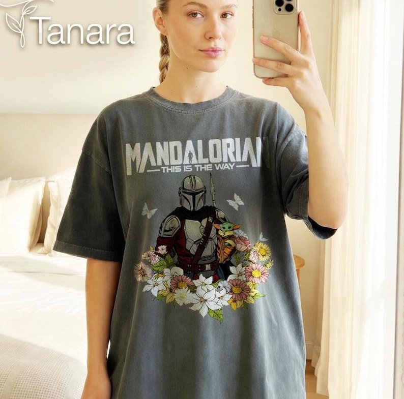 Puede incluir: Camiseta gris con el texto "MANDALORIAN - THIS IS THE WAY" y una ilustraci&oacute;n del Mandaloriano, Grogu y flores. La camiseta tiene mangas cortas y un corte holgado. La marca "Tanara" est&aacute; en la esquina superior izquierda.