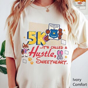 May include: Ivory-colored t-shirt with a colorful graphic design. The design includes the text "5K... It's Called a Hustle, Sweetheart" with cartoon characters and a badge that says "Walt Disney World Springtime Surprise 2026".