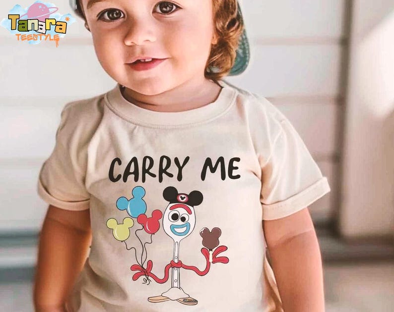 K&ouml;nnte beinhalten: Ein beiges T-Shirt mit den Worten "CARRY ME" in Schwarz. Unter dem Text befindet sich eine Zeichentrickfigur, die Luftballons h&auml;lt. Die Ballons sind blau, gelb und rot. Das T-Shirt ist f&uuml;r Kinder.