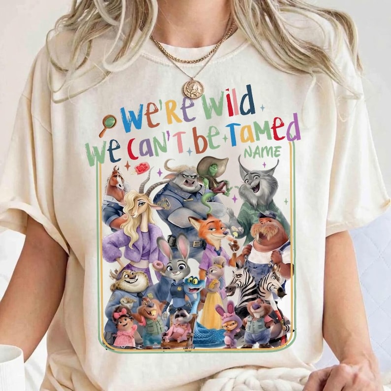 May include: Cream-colored t-shirt featuring a colorful graphic of animated animal characters. The text "WE'RE WILD WE CAN'T BE TAMED" is displayed above the characters. A space is provided for a name. The shirt has short sleeves and a crew neck.