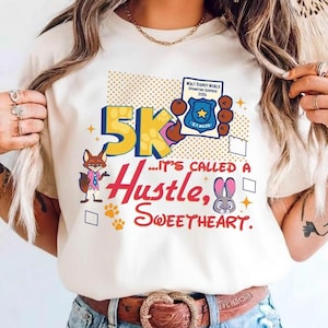May include: Cream-colored t-shirt with a colorful graphic design. The design includes the text "5K... It's Called a Hustle, Sweetheart" with cartoon characters and a badge. The shirt has a crew neck and short sleeves.