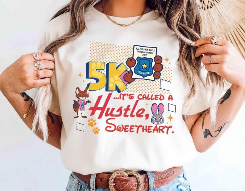 May include: Cream-colored t-shirt with a colorful graphic design. The design includes the text "5K... It's Called a Hustle, Sweetheart" with cartoon characters and a badge. The shirt has a crew neck and short sleeves.