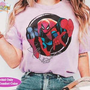 May include: A light purple t-shirt featuring a graphic of Spider-Man in a black circle. The superhero is depicted in his classic red and blue suit, with a web and chain detail. The text "Orchid Only With Comfort Color" is at the bottom.