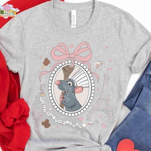 May include: A light gray t-shirt featuring a cartoon rat in a decorative frame with a pink bow, Eiffel Tower, and hearts. The text "I Love Paris" is also present. The shirt is paired with blue jeans and a red jacket.