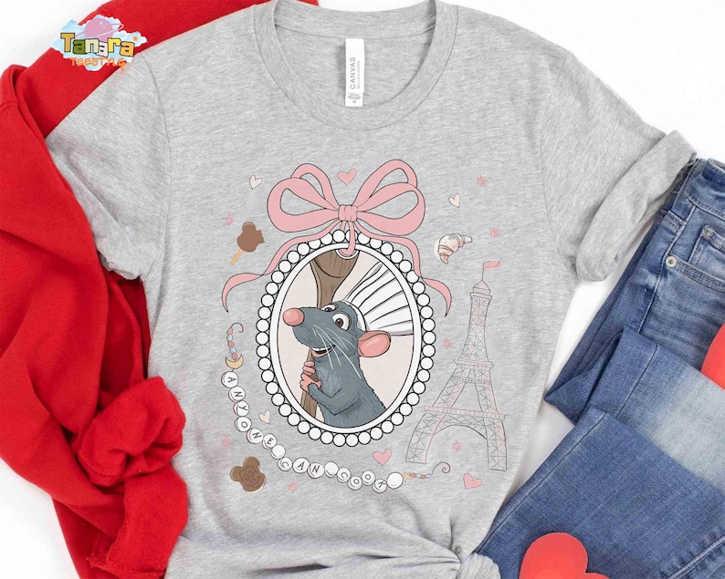 May include: A light gray t-shirt featuring a cartoon rat in a decorative frame with a pink bow, Eiffel Tower, and hearts. The text "I Love Paris" is also present. The shirt is paired with blue jeans and a red jacket.