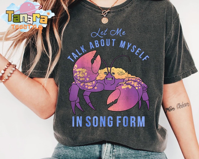 Puede incluir: Camiseta gris oscuro con una ilustraci&oacute;n de cangrejo morado con pinzas rosas y el texto "Let Me Talk About Myself In Song Form". La camiseta tiene un aspecto vintage descolorido.