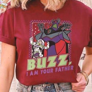 May include: Burgundy t-shirt with a graphic of Buzz Lightyear and Zurg from Toy Story. The design includes the text "BUZZ, I AM YOUR FATHER" in green and purple. The shirt has short sleeves and a crew neck.
