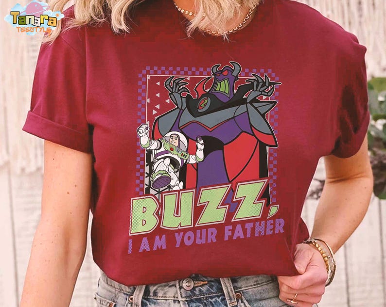 May include: Burgundy t-shirt with a graphic of Buzz Lightyear and Zurg from Toy Story. The design includes the text "BUZZ, I AM YOUR FATHER" in green and purple. The shirt has short sleeves and a crew neck.