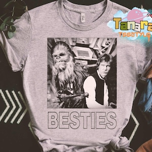 May include: A grey t-shirt with a black and white image of Chewbacca and Han Solo from Star Wars. The word "BESTIES" is printed in black block letters below the image. The shirt has a crew neck and short sleeves.
