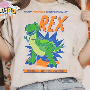 Puede incluir: Camiseta beige con un dinosaurio de dibujos animados verde y la palabra naranja "REX". El texto dice "THE MOST TERRIFYING DINOSAUR WHO EVER LIVED!" y "AVAILABLE AT TOY STORES EVERYWHERE!" en la camiseta.