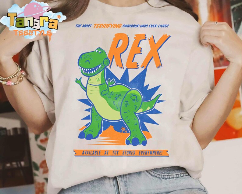 Puede incluir: Camiseta beige con un dinosaurio de dibujos animados verde y la palabra naranja "REX". El texto dice "THE MOST TERRIFYING DINOSAUR WHO EVER LIVED!" y "AVAILABLE AT TOY STORES EVERYWHERE!" en la camiseta.
