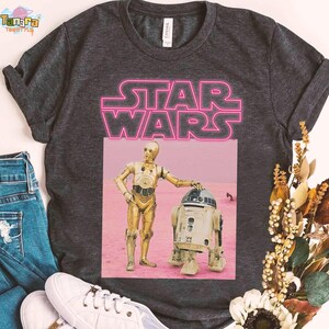 May include: Dark gray t-shirt featuring a pink neon "STAR WARS" graphic. Below the text is a rectangular image of C-3PO and R2-D2 on a pink background. The shirt is displayed with blue jeans and white sneakers.