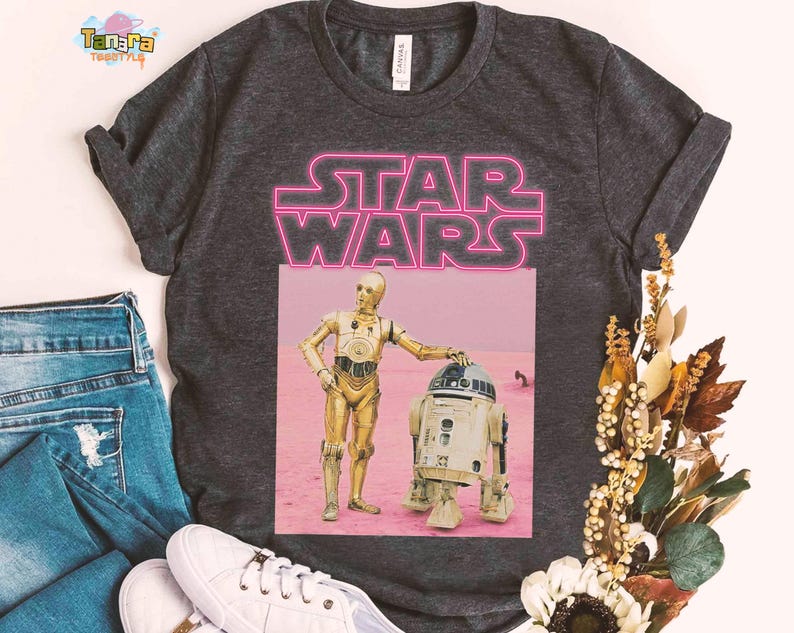 May include: Dark gray t-shirt featuring a pink neon "STAR WARS" graphic. Below the text is a rectangular image of C-3PO and R2-D2 on a pink background. The shirt is displayed with blue jeans and white sneakers.