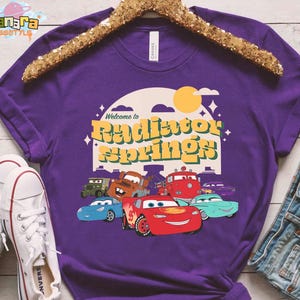 Pode incluir: Uma camiseta roxa com a frase "Welcome to Radiator Springs" e personagens de desenhos animados de carros. O design inclui um sol amarelo e ilustra&ccedil;&otilde;es de carros. A camiseta &eacute; exibida com t&ecirc;nis brancos e shorts jeans.