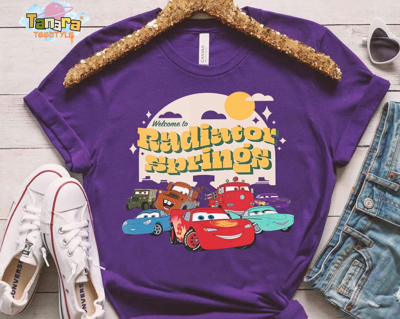 Pode incluir: Uma camiseta roxa com a frase "Welcome to Radiator Springs" e personagens de desenhos animados de carros. O design inclui um sol amarelo e ilustra&ccedil;&otilde;es de carros. A camiseta &eacute; exibida com t&ecirc;nis brancos e shorts jeans.