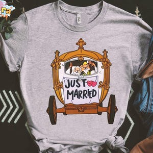 May include: A gray t-shirt featuring a cartoon carriage with the words "JUST MARRIED" on a banner. The carriage has two cartoon characters inside, and two pink hearts are on the banner. The shirt has a crew neck.