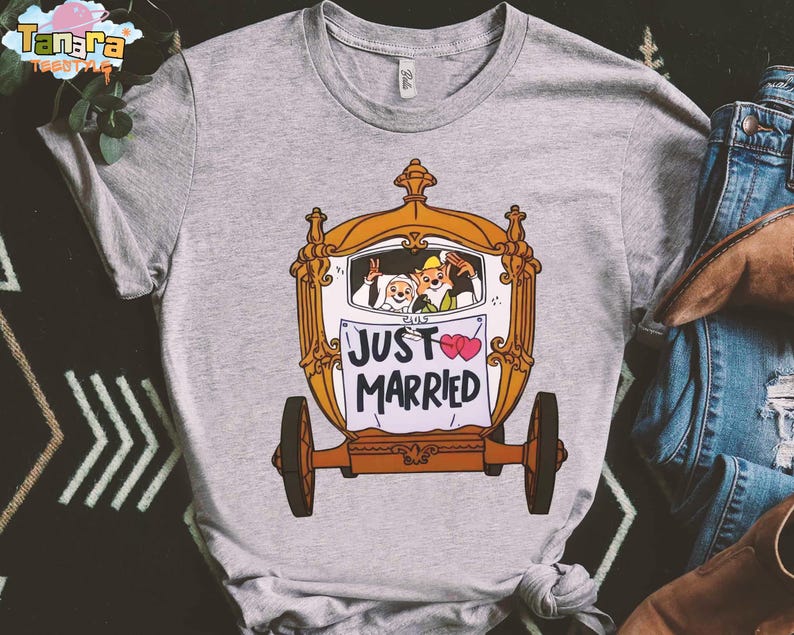 May include: A gray t-shirt featuring a cartoon carriage with the words "JUST MARRIED" on a banner. The carriage has two cartoon characters inside, and two pink hearts are on the banner. The shirt has a crew neck.