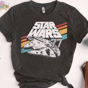 Puede incluir: Camiseta gris oscuro con un gráfico del Halcón Milenario y las palabras "STAR WARS 77" en blanco. El diseño incluye rayas retro en rojo, naranja, amarillo y turquesa. La camiseta es de cuello redondo.