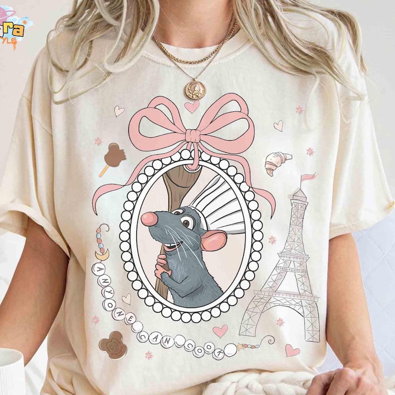 May include: Cream-colored t-shirt featuring a cartoon rat chef inside an oval frame with a pink bow. The design includes the Eiffel Tower, hearts, and the text "ANYONE CAN COOK".