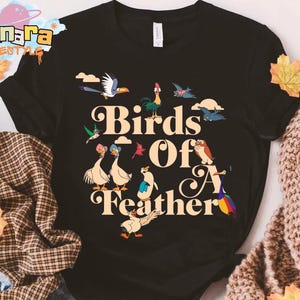 May include: Black t-shirt with the text "Birds Of A Feather" in a whimsical font, accompanied by cartoon illustrations of various birds. The design includes geese, a rooster, and other colorful birds.