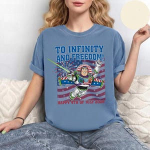 May include: A light blue t-shirt featuring Buzz Lightyear with the text "TO INFINITY AND FREEDOM!" and "HAPPY 4TH OF JULY 2026" in red. The design includes a red, white, and blue American flag and fireworks.