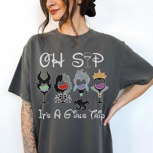 May include: Dark gray t-shirt with the text "OH SIP It's A Girls Trip". The shirt features four cartoon wine glasses, each with a different villain-inspired design and color scheme. The design includes a crown, horns, and other details.
