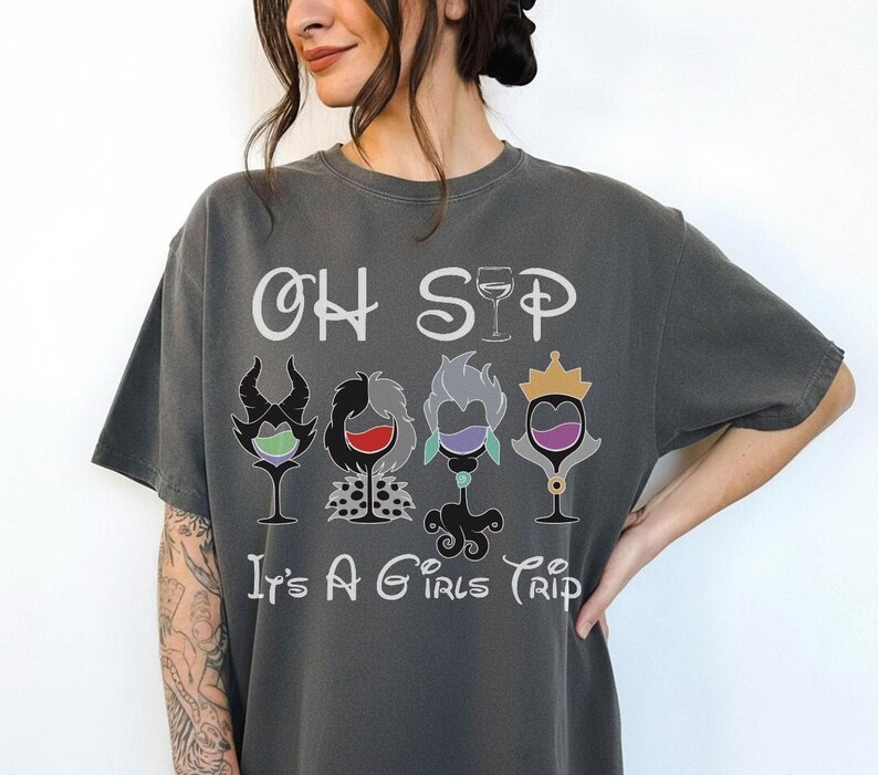May include: Dark gray t-shirt with the text "OH SIP It's A Girls Trip". The shirt features four cartoon wine glasses, each with a different villain-inspired design and color scheme. The design includes a crown, horns, and other details.