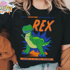 Puede incluir: Camiseta negra con un dinosaurio de dibujos animados llamado Rex. El dinosaurio verde tiene una expresi&oacute;n amigable y est&aacute; sobre un fondo azul y naranja. El texto en la camiseta dice "THE MOST TERRIFYING DINOSAUR WHO EVER LIVED!" y "AVAILABLE AT TOY STORES EVERYWHERE!"