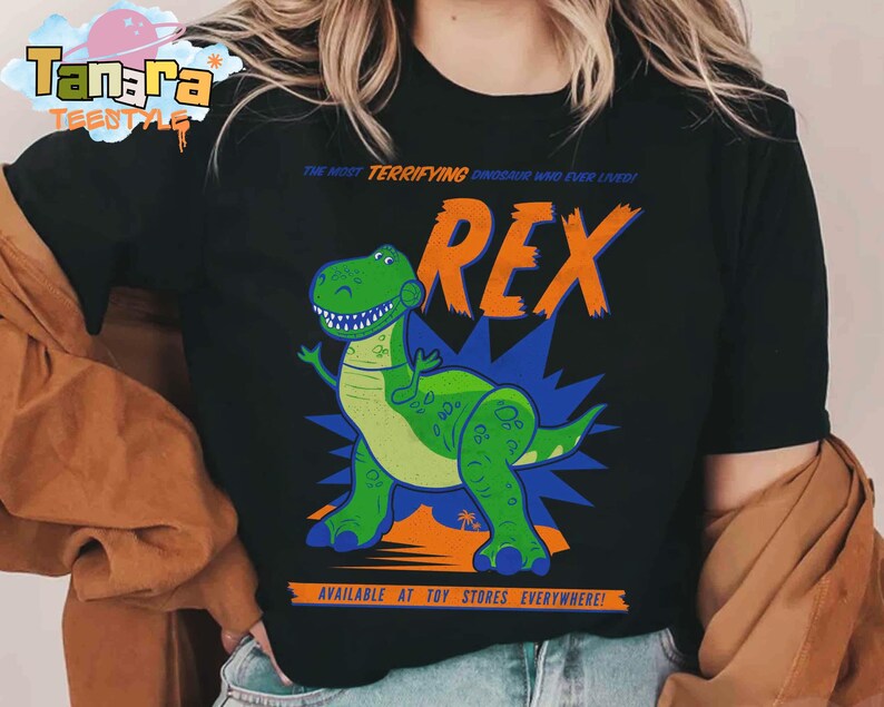 Puede incluir: Camiseta negra con un dinosaurio de dibujos animados llamado Rex. El dinosaurio verde tiene una expresi&oacute;n amigable y est&aacute; sobre un fondo azul y naranja. El texto en la camiseta dice "THE MOST TERRIFYING DINOSAUR WHO EVER LIVED!" y "AVAILABLE AT TOY STORES EVERYWHERE!"