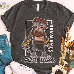 May include: Dark grey t-shirt with a Babu Frik cartoon character from Star Wars. The design includes the text "STAR WARS" and "BABU FRIK" in white. The shirt is styled with blue jeans and white trainers.