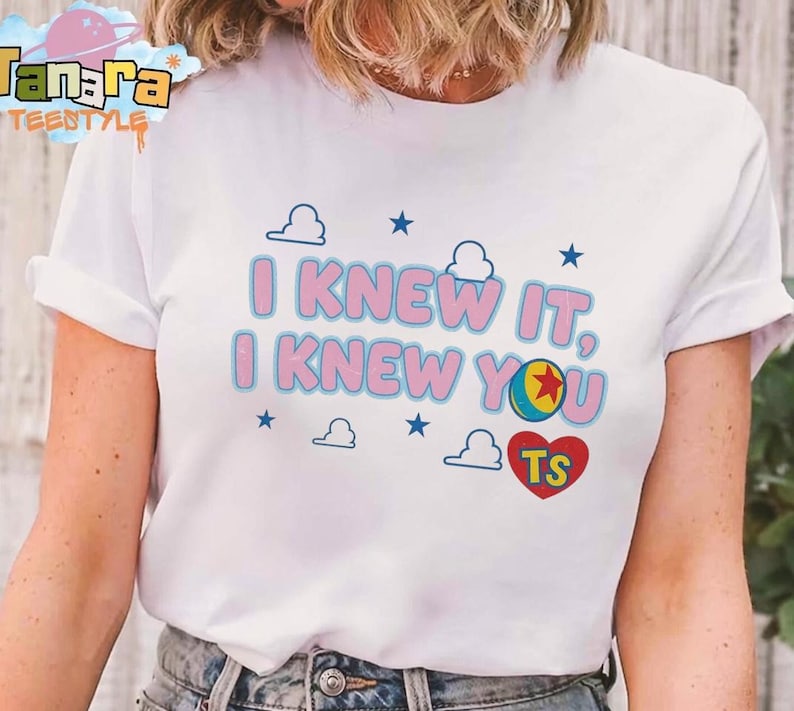 May include: A white short-sleeved t-shirt featuring the pink text I KNEW IT, I KNEW YOU with a blue outline. The design includes small blue stars, white cloud outlines, a colorful ball with a red star, and a red heart containing the letters TS.