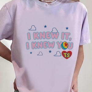 K&ouml;nnte beinhalten: Ein hellviolettes Kurzarm-T-Shirt mit dem Text I KNEW IT, I KNEW YOU in Pink mit blauen Umrandungen. Die Grafik enth&auml;lt einen bunten Ball, ein rotes Herz mit den Buchstaben TS sowie mehrere kleine blaue Wolken und Sterne im Hintergrund.