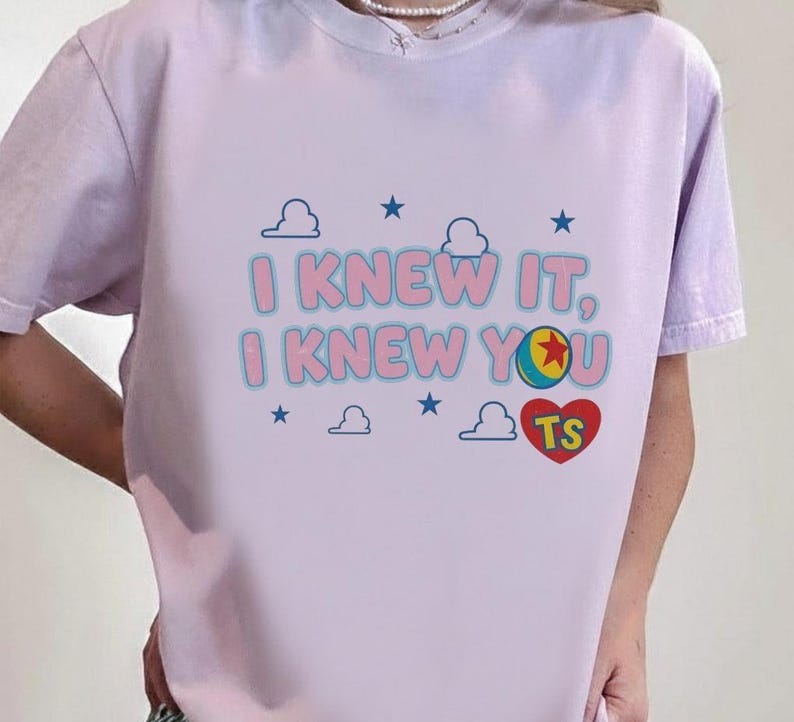 K&ouml;nnte beinhalten: Ein hellviolettes Kurzarm-T-Shirt mit dem Text I KNEW IT, I KNEW YOU in Pink mit blauen Umrandungen. Die Grafik enth&auml;lt einen bunten Ball, ein rotes Herz mit den Buchstaben TS sowie mehrere kleine blaue Wolken und Sterne im Hintergrund.