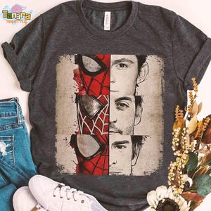 Puede incluir: Camiseta gris oscuro con un gr&aacute;fico de la m&aacute;scara de Spider-Man y los rostros de tres actores. El dise&ntilde;o incluye elementos rojos, blancos y negros. La camiseta se muestra con vaqueros azules y zapatillas blancas.