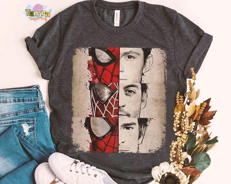 Puede incluir: Camiseta gris oscuro con un gr&aacute;fico de la m&aacute;scara de Spider-Man y los rostros de tres actores. El dise&ntilde;o incluye elementos rojos, blancos y negros. La camiseta se muestra con vaqueros azules y zapatillas blancas.
