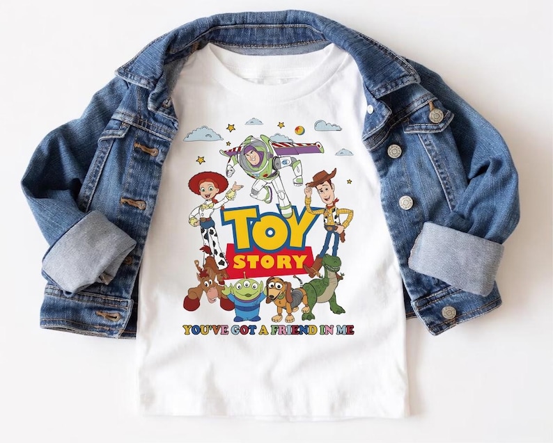 May include: White t-shirt featuring the Toy Story characters Buzz Lightyear, Woody, Jessie, and others. The shirt has the words "TOY STORY" in yellow and red, with the phrase "YOU'VE GOT A FRIEND IN ME" below. A blue denim jacket is draped over the shirt.