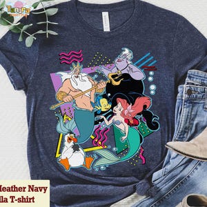 May include: Heather navy Bella T-shirt with a vibrant graphic of animated film characters. The design includes a mermaid, a sea witch, and other sea creatures. The shirt is paired with blue jeans and a boot.