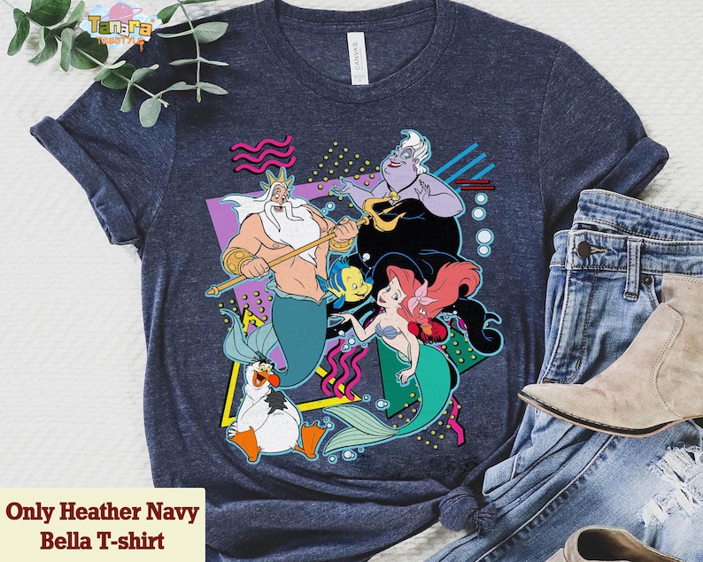 May include: Heather navy Bella T-shirt with a vibrant graphic of animated film characters. The design includes a mermaid, a sea witch, and other sea creatures. The shirt is paired with blue jeans and a boot.