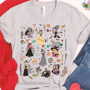 May include: A light gray t-shirt featuring a colorful Halloween-themed graphic. The design includes illustrations of witches, pumpkins, bats, and other spooky elements. The shirt is paired with blue jeans and a red sweatshirt.